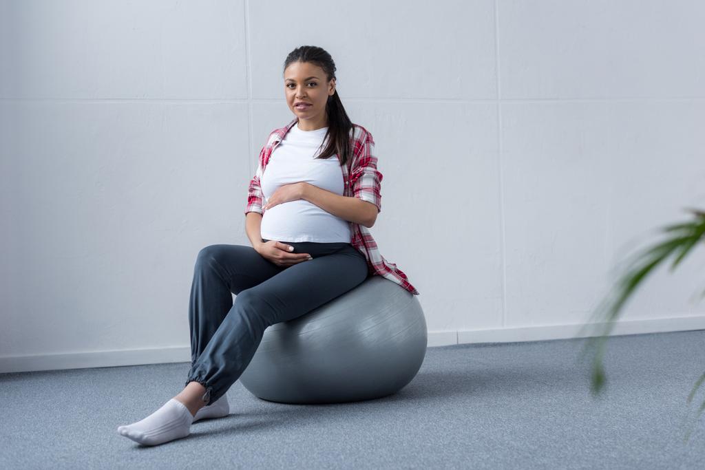 フィット ボールの上に座って陽気なアフリカ系アメリカ人の妊娠中の女性 - 写真・画像