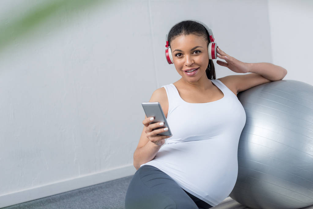 アフリカ系アメリカ人の妊娠中の女性フィット ボールと一緒に座っている間スマート フォンとヘッドフォンの音楽を聴く - 写真・画像
