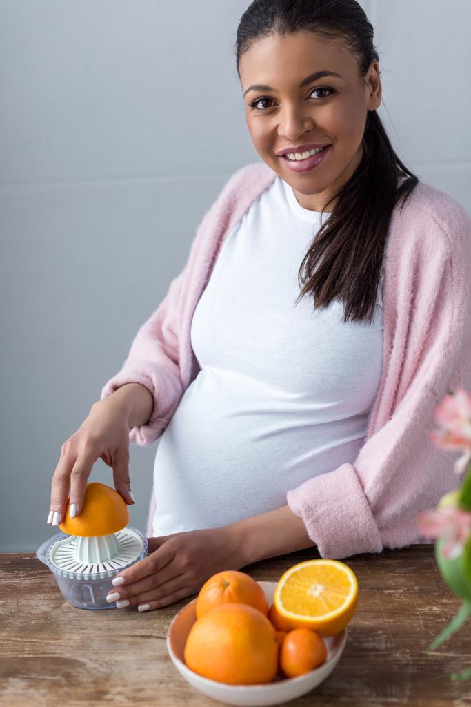 bela mulher grávida afro-americana sorridente fazendo suco de laranja com espremedor
 - Foto, Imagem