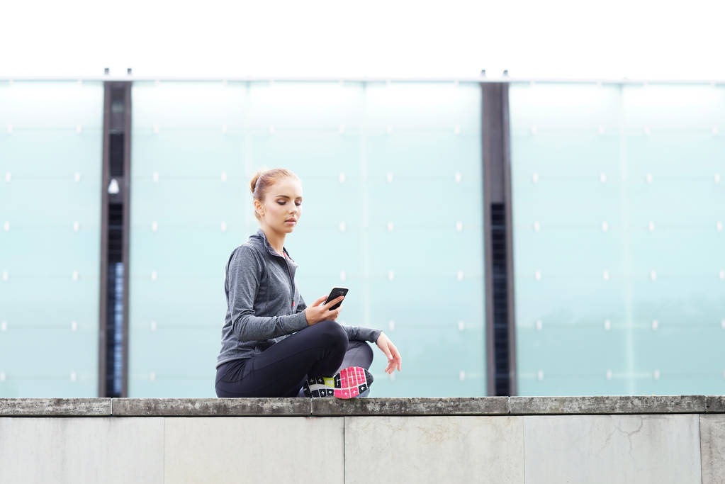 Femme jeune, en forme et sportive assise sur la frontière en béton avec téléphone. Fitness, sport, jogging urbain et mode de vie sain concept
. - Photo, image