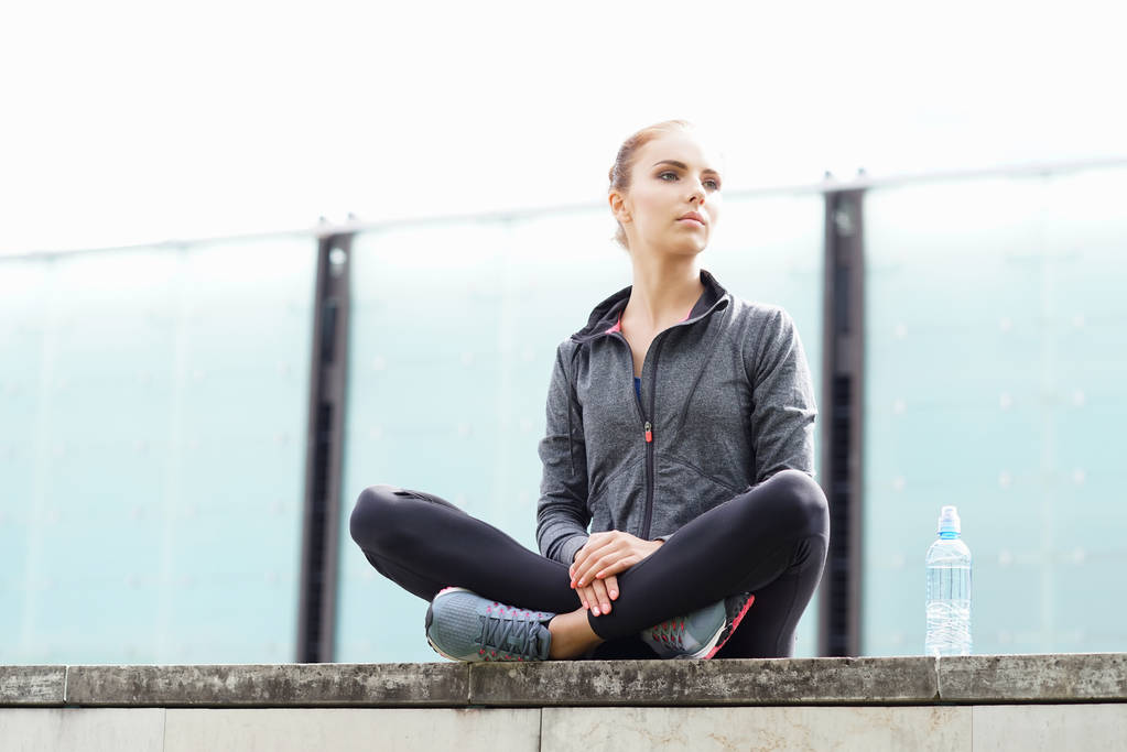 Молодая, стройная и спортивная женщина, сидящая на бетонной границе. Фитнес, спорт, бег трусцой и концепция здорового образа жизни
. - Фото, изображение