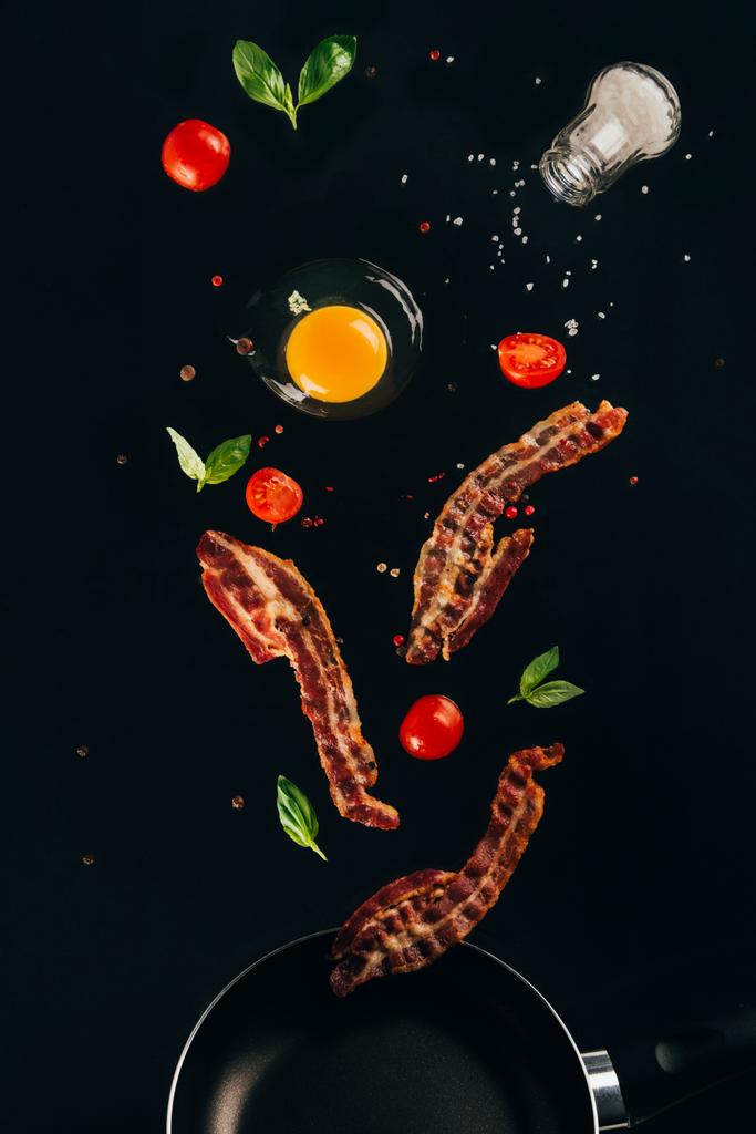κλείνω πάνω θέα από κομμάτια του μπέικον, τα ντοματίνια και πρώτης κρόκο αυγού που πέφτουν στο τηγάνι που απομονώνονται σε μαύρο - Φωτογραφία, εικόνα