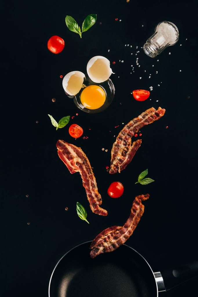 close up view of pieces of bacon, cherry tomatoes and raw egg yolk falling on frying pan isolated on black - Photo, Image