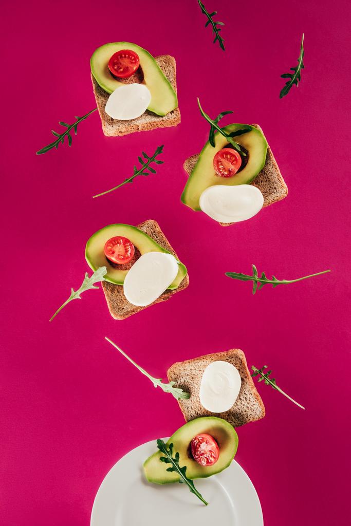 close up view of toasts, avocado pieces, mozzarella cheese and cherry tomatoes falling on plate isolated on pink - Photo, Image