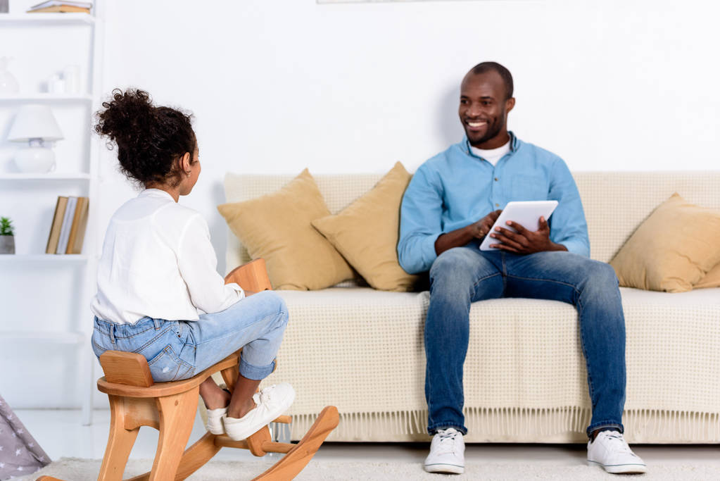 Afrikanisch-amerikanischer Vater mit Tablet sieht Tochter zu Hause auf Schaukelpferd sitzen - Foto, Bild