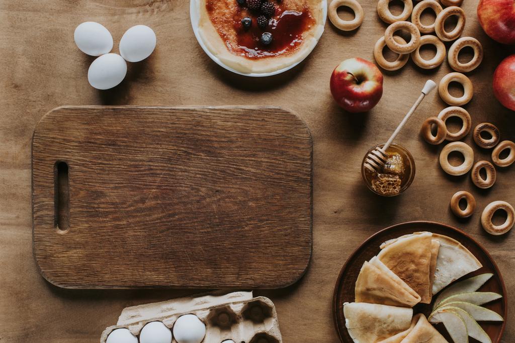 Blick von oben auf leckere hausgemachte Pfannkuchen, Eier und Eierschachtel, Holzschneidebrett, Äpfel, Honig und Bagels auf dem Tisch - Foto, Bild