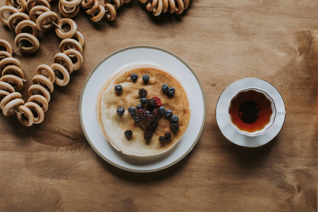 Blick von oben auf leckere Pfannkuchen mit Beeren, Tee und Bagels auf Holztisch  - Foto, Bild