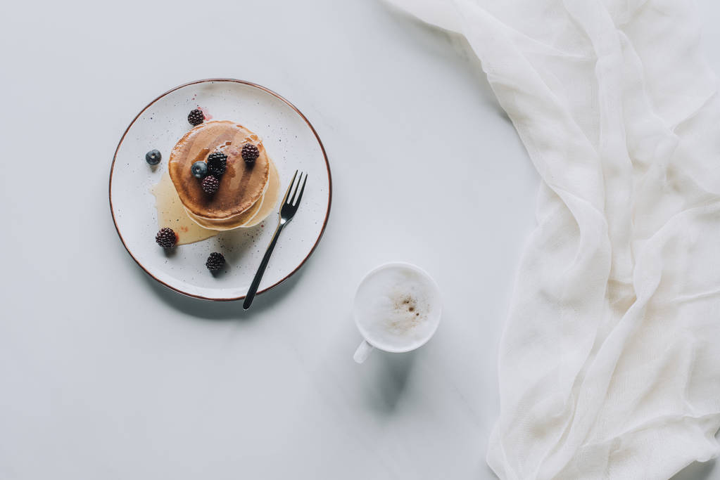 Blick von oben auf Tasse Cappuccino und süße leckere Pfannkuchen mit Beeren und Honig auf grau - Foto, Bild