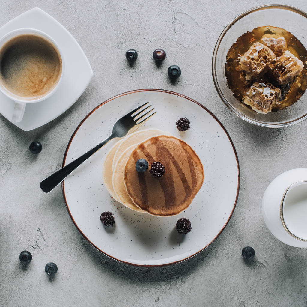 bovenaanzicht van heerlijke pannenkoeken met jam en bessen, koffie met melk en honing op grijs - Foto, afbeelding