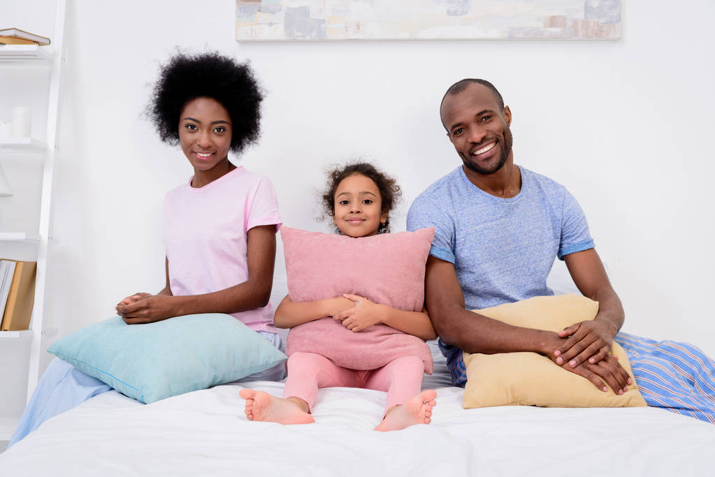 uśmiechający się african american rodziców i córka siedzi z poduszkami i patrząc na kamery - Zdjęcie, obraz