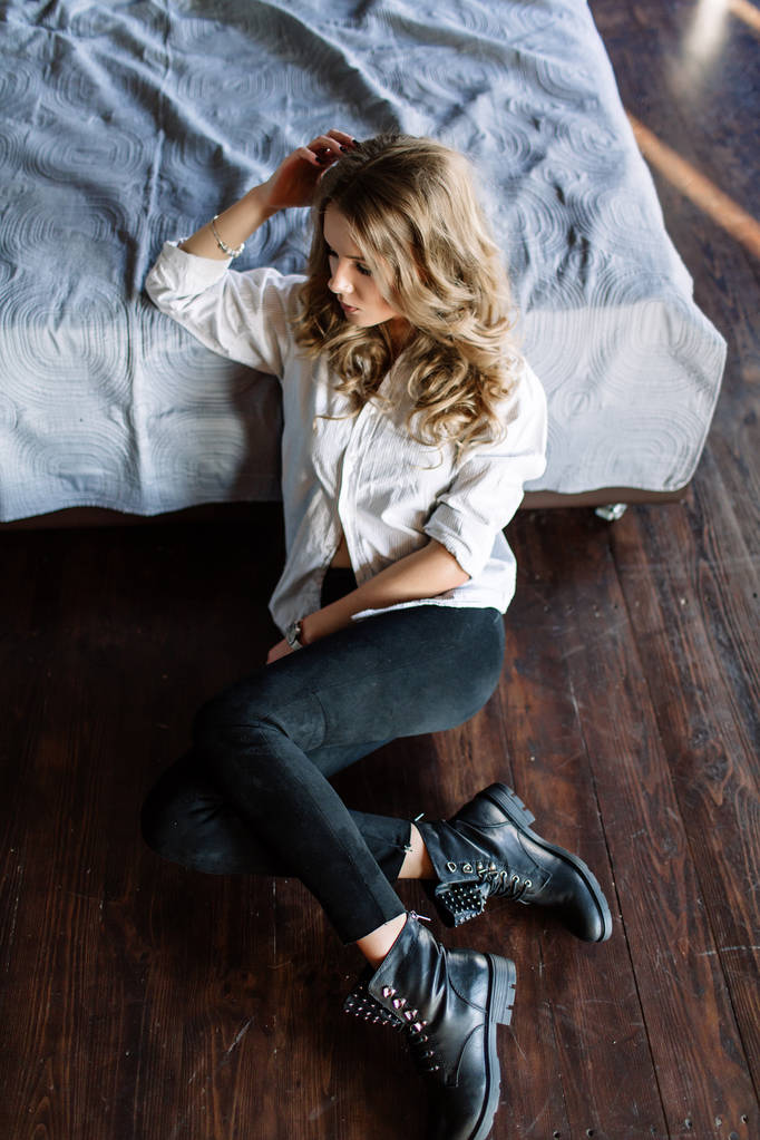 Une fille aux longs cheveux blonds vêtus de jeans noirs, une chemise rayée et des chaussures noires assises sur le lit avec des draps bleus. Tenue casual à la mode
. - Photo, image