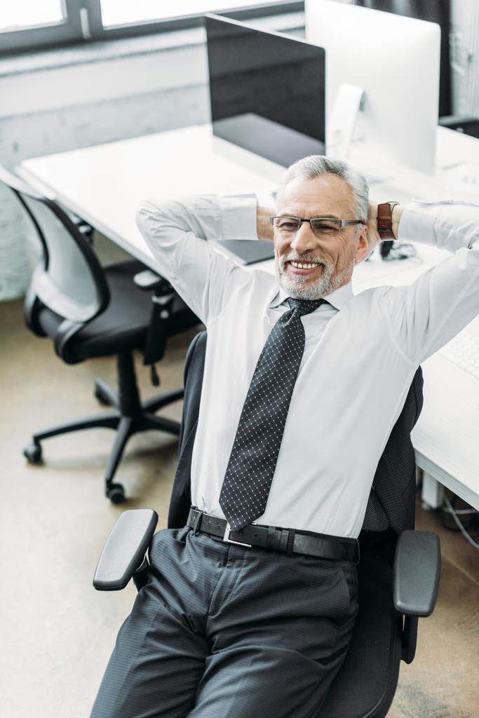 Portret van lachende senior zakenman met handen achter hoofd in kantoor - Foto, afbeelding