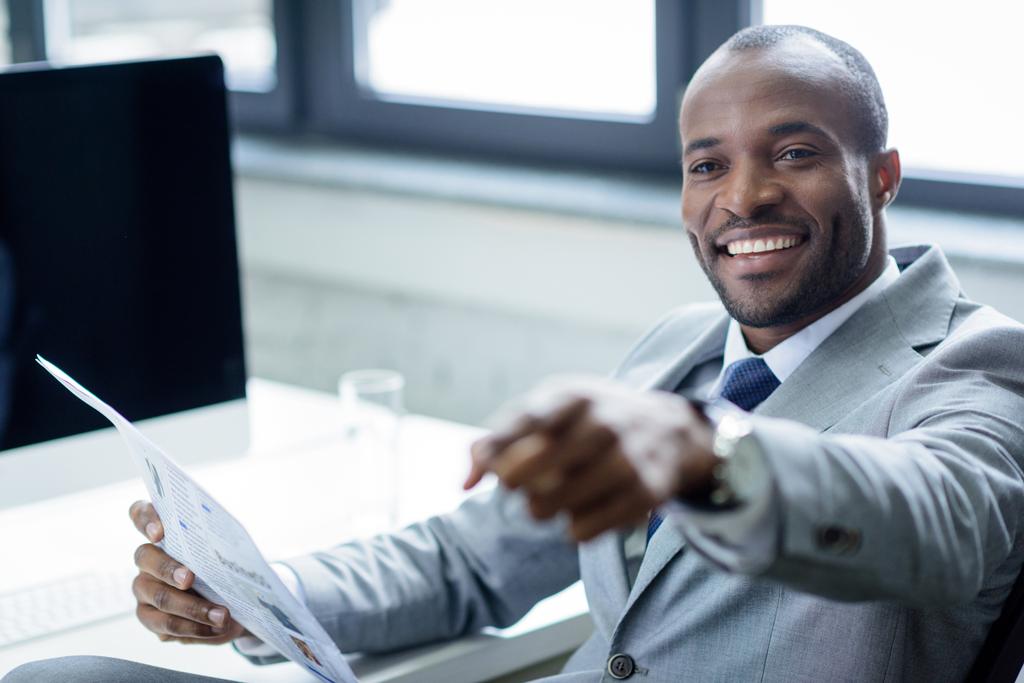 sonriente hombre de negocios afroamericano con periódico apuntando hacia el lugar de trabajo
 - Foto, imagen