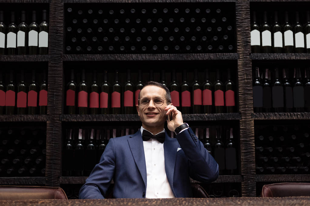 hombre guapo en traje elegante hablando por teléfono en el restaurante frente a los estantes de almacenamiento de vino
 - Foto, Imagen