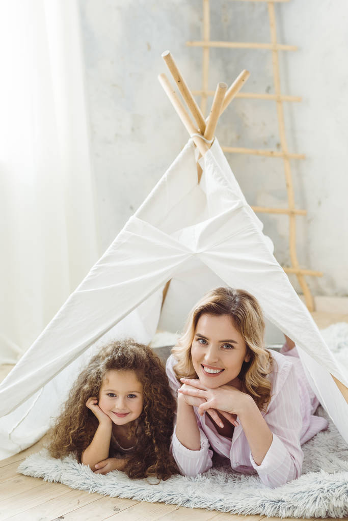beautiful cheerful mother and curly daughter lying in kid wigwam - Photo, Image