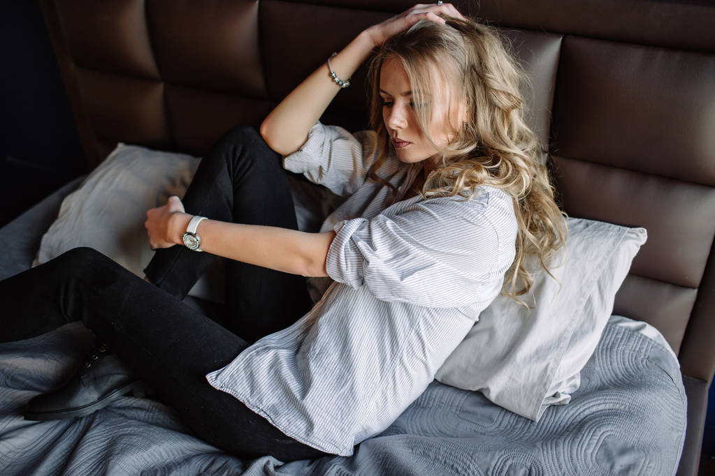 A girl with long blond hair dressed in black jeans, a striped shirt and black shoes sitting on the bed with blue linens. Fashionable casual outfit. - Photo, Image