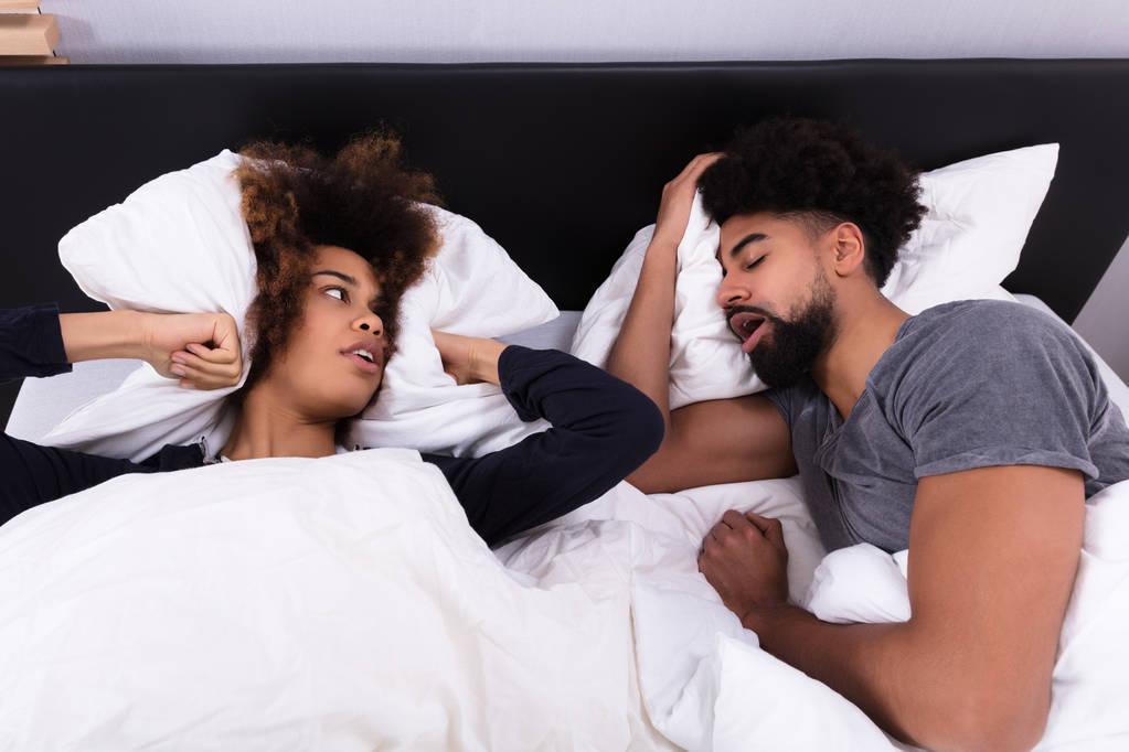 Повышенный вид молодой женщины, закрывающей уши подушкой, пока мужчина храпит в постели
 - Фото, изображение