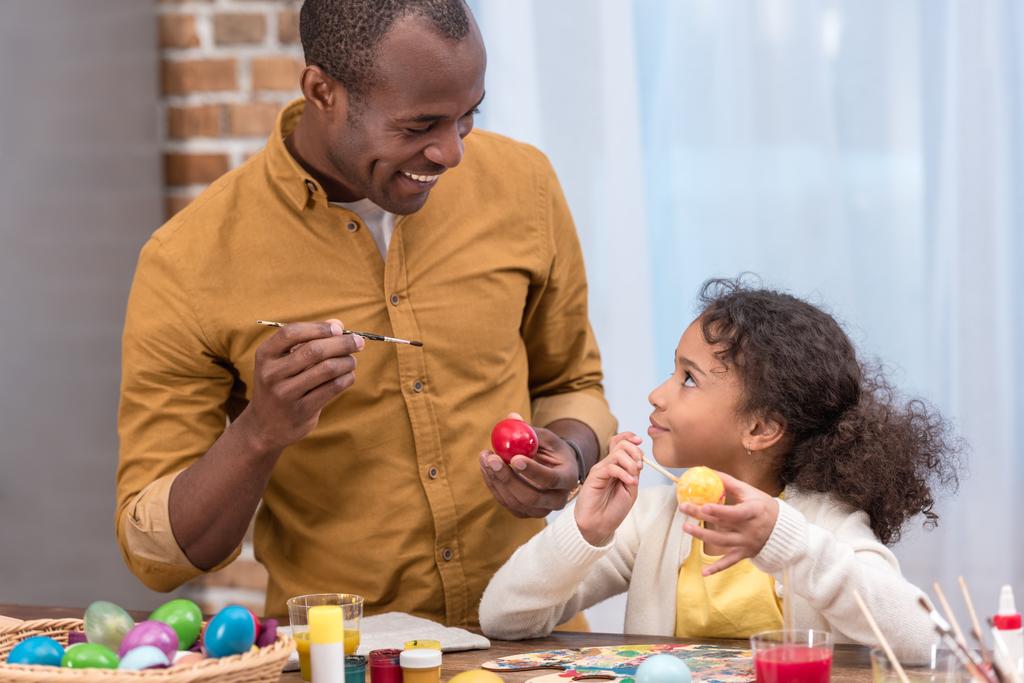 Африканский американец отец и дочь рисуют пасхальные яйца и смотрят друг на друга
 - Фото, изображение
