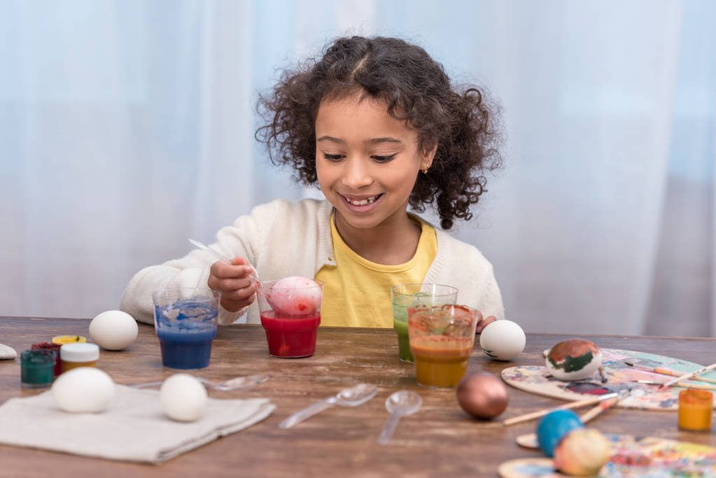 fröhliches afrikanisch-amerikanisches Kind bemalt Ostereier in Gläsern mit Farben - Foto, Bild