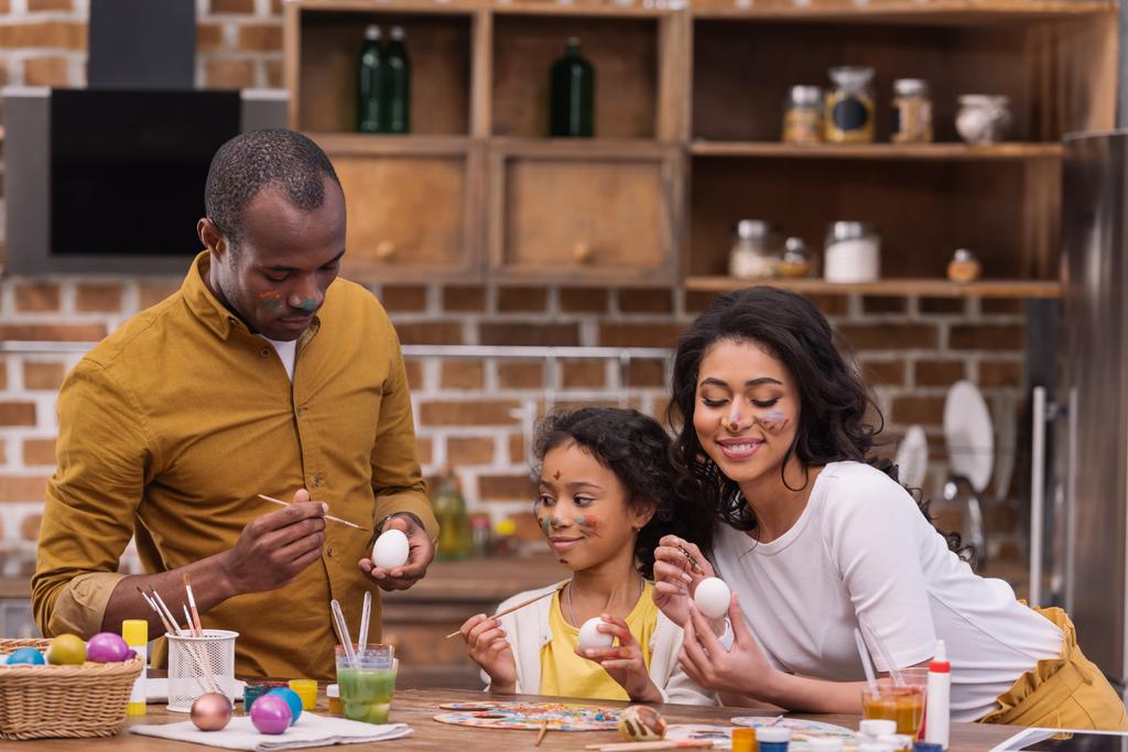 χαμογελώντας αφρικανικές αμερικανικές γονείς και κόρη ζωγραφική αυγά του Πάσχα μαζί στην κουζίνα - Φωτογραφία, εικόνα