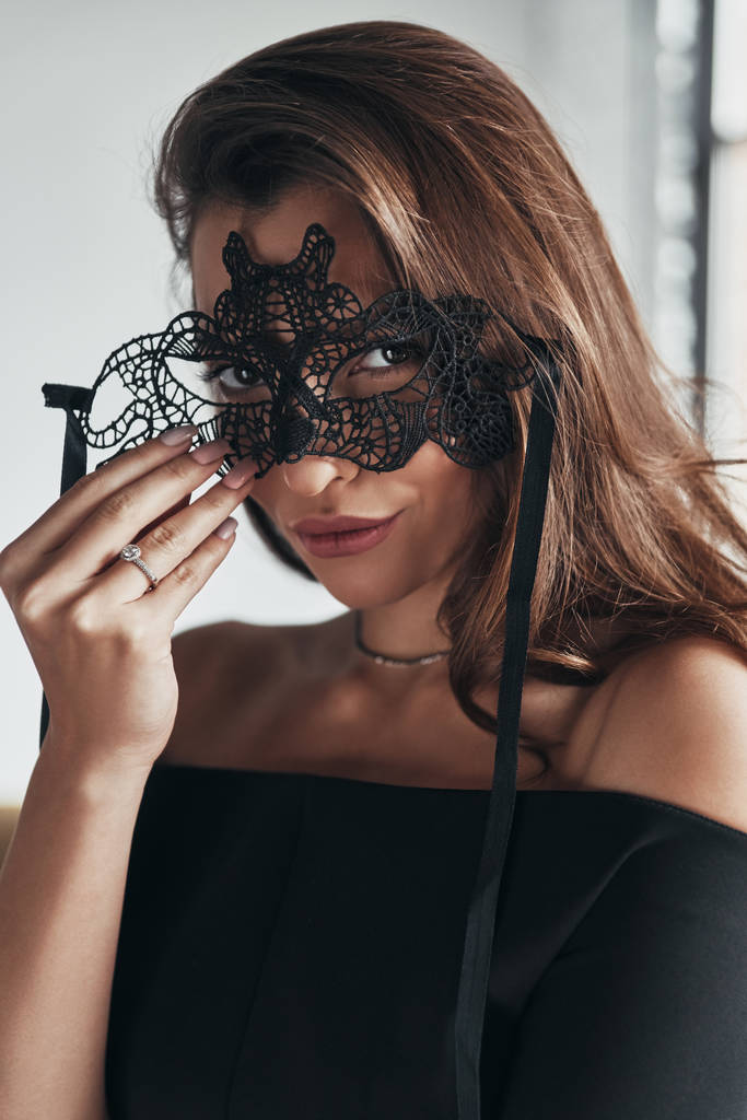 Nádherná žena držící černé karnevalové masky na obličej a při pohledu na fotoaparát - Fotografie, Obrázek