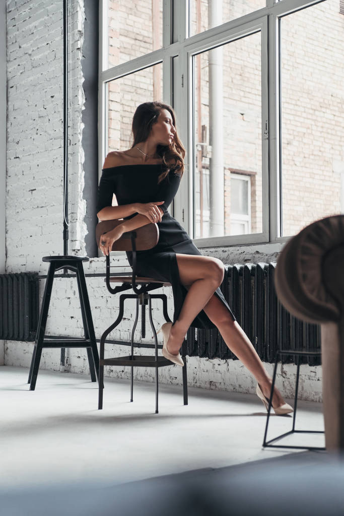 brunetka Kobieta w eleganckiej czarnej sukni siedzi na krześle w domu - Zdjęcie, obraz