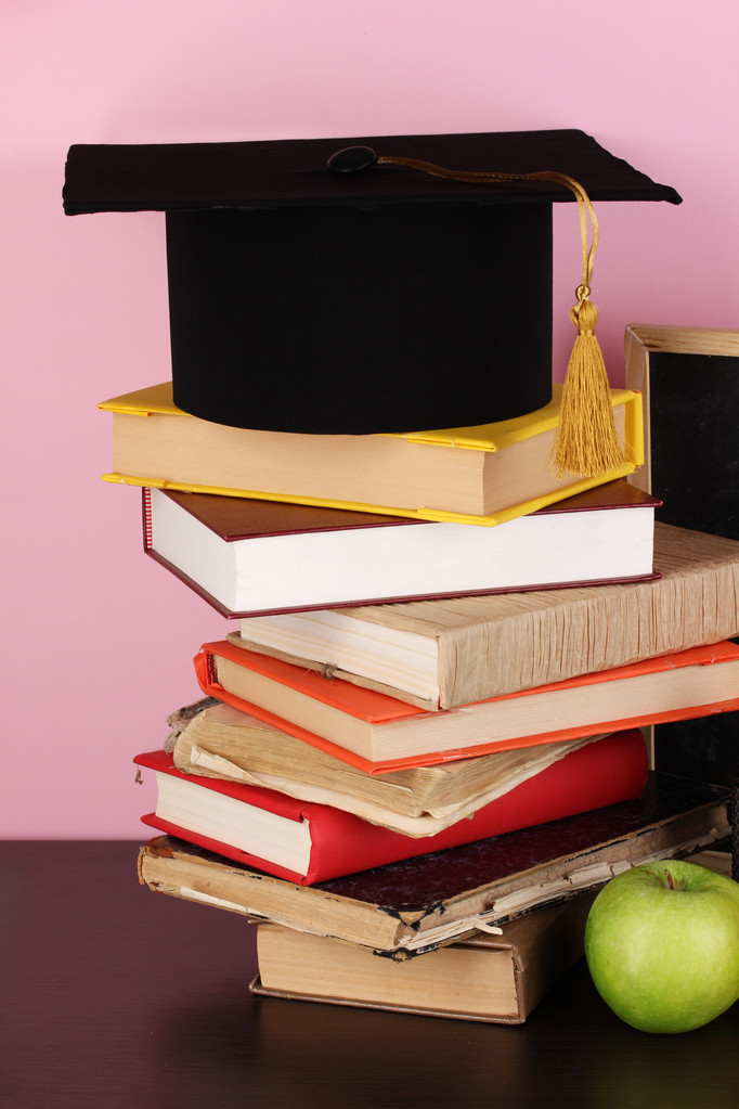 Bücher und Magistermütze gegen Schultafel auf Holztisch auf rosa Hintergrund - Foto, Bild