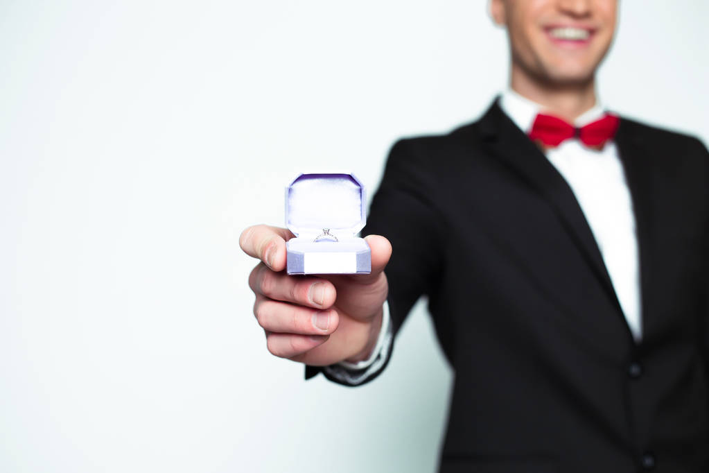 Yakışıklı gülümseyen modern zarif insan resmi siyah takım elbise ve kırmızı papyon bir nişan yüzüğü ile bir kutu koruyor ve kamera gösterir. - Fotoğraf, Görsel