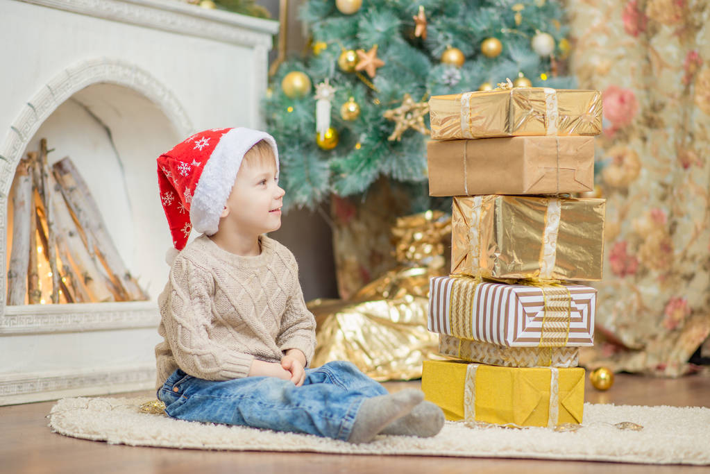 um menino em um boné vermelho senta-se perto de uma árvore de Natal com um presente
 - Foto, Imagem