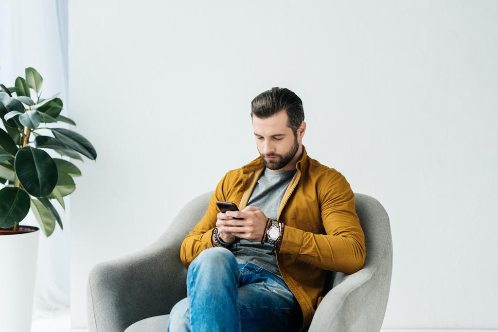 knappe man in leunstoel zitten en het gebruik van smartphone - Foto, afbeelding