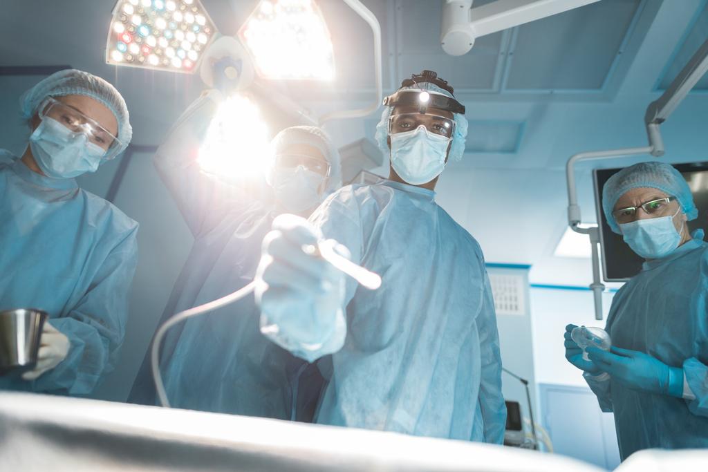visão inferior de cirurgiões multiculturais olhando para a câmera durante a cirurgia
 - Foto, Imagem