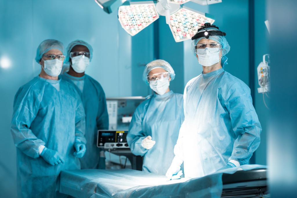 neljä monikulttuurista lääkäriä katselemassa kameraa leikkaussalissa
 - Valokuva, kuva
