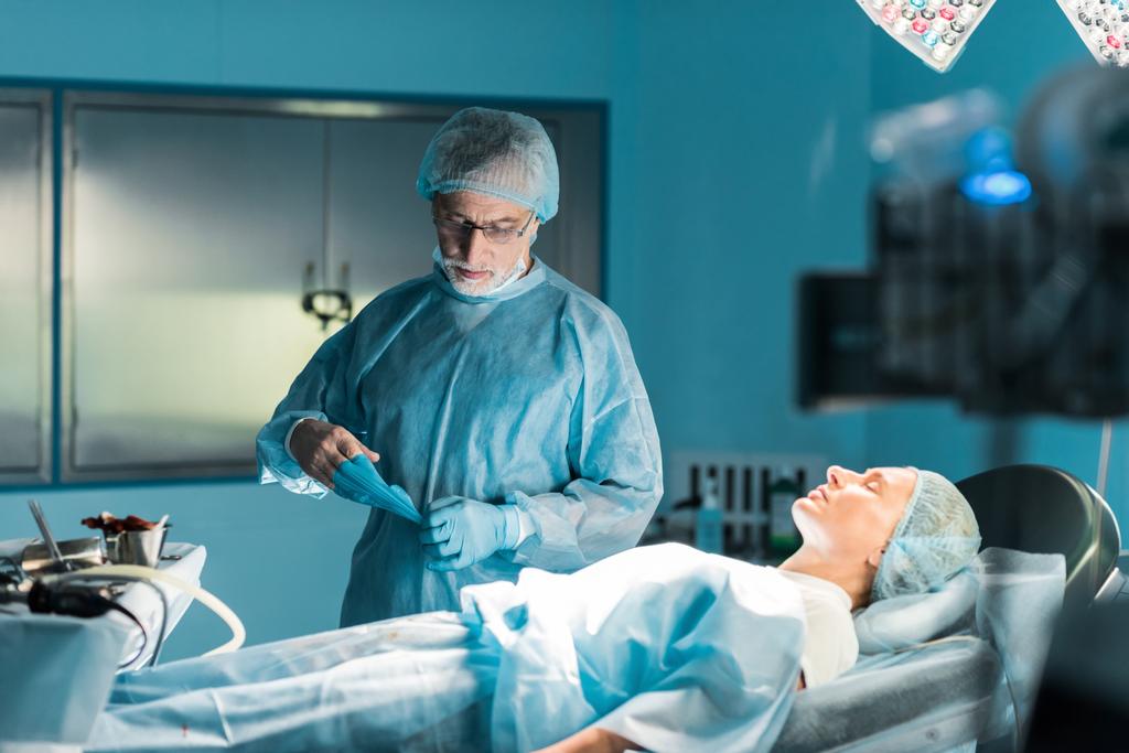 Врач снимает медицинские перчатки рядом с пациентом в операционной
 - Фото, изображение