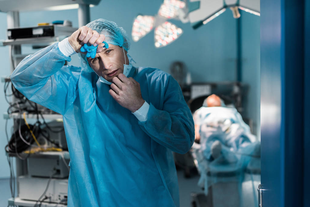 усталый врач снимает медицинскую маску и трогает лоб в операционной
 - Фото, изображение