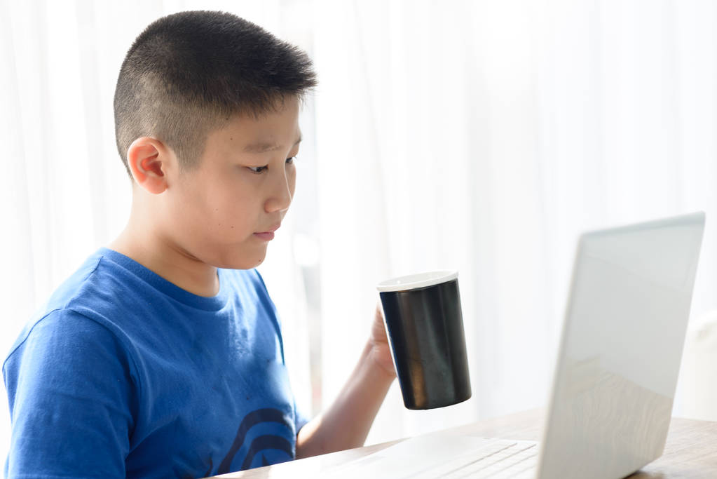 Ασίας αγόρι κρατώντας μαύρα Κύπελλο ενώ χρησιμοποιείτε tablet στο σπίτι. - Φωτογραφία, εικόνα