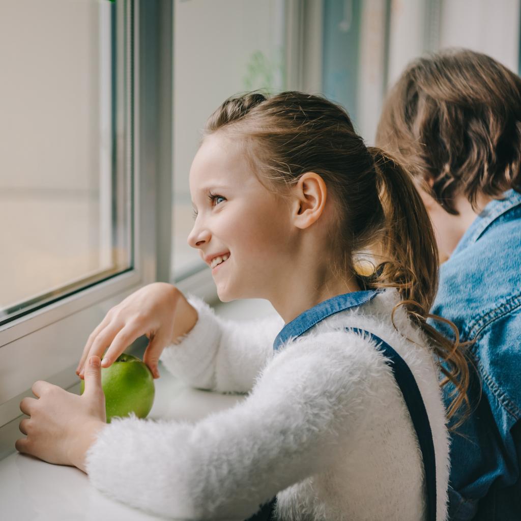 beautiful schoolgirl with fresh apple looking through window at classroom - Photo, Image