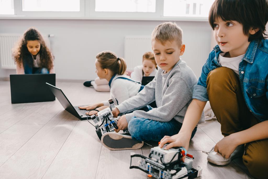 εστιασμένη παιδιά προγραμματισμός ρομπότ με φορητούς υπολογιστές, ενώ κάθεται στο πάτωμα, στέλεχος εκπαίδευσης έννοια - Φωτογραφία, εικόνα