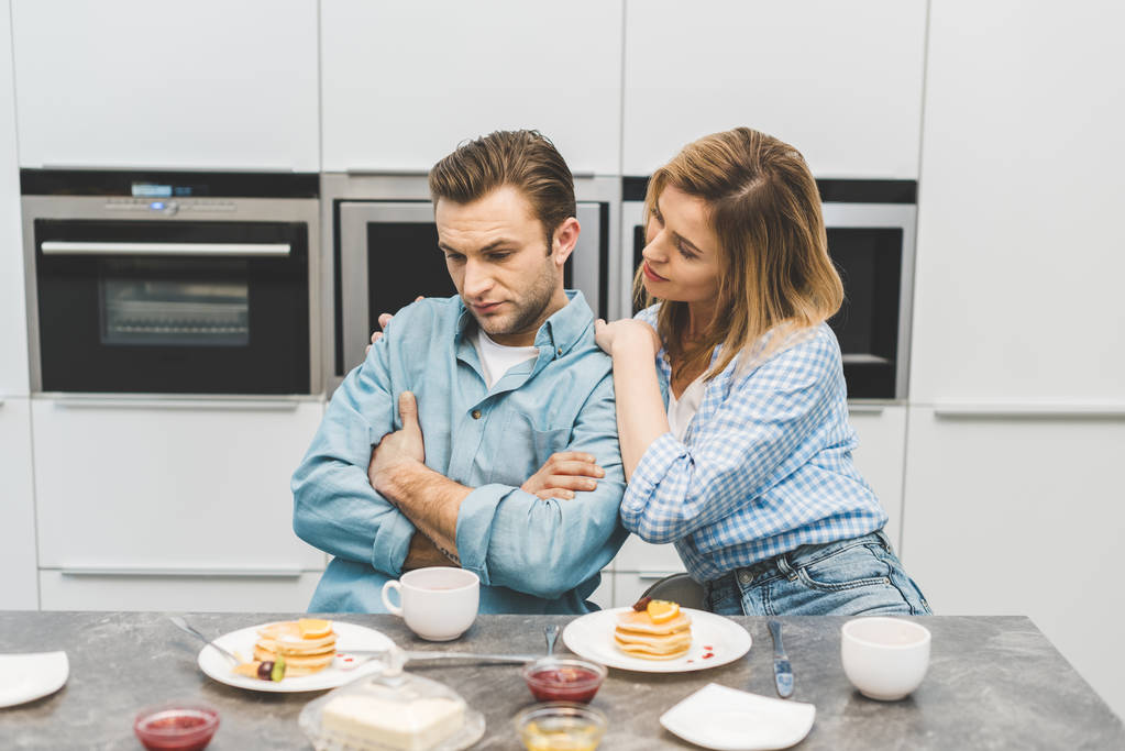 vrouw knuffelen boos man tijdens het ontbijt thuis - Foto, afbeelding