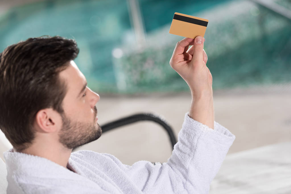 jovem bonito em roupão segurando cartão de crédito no centro de spa
  - Foto, Imagem