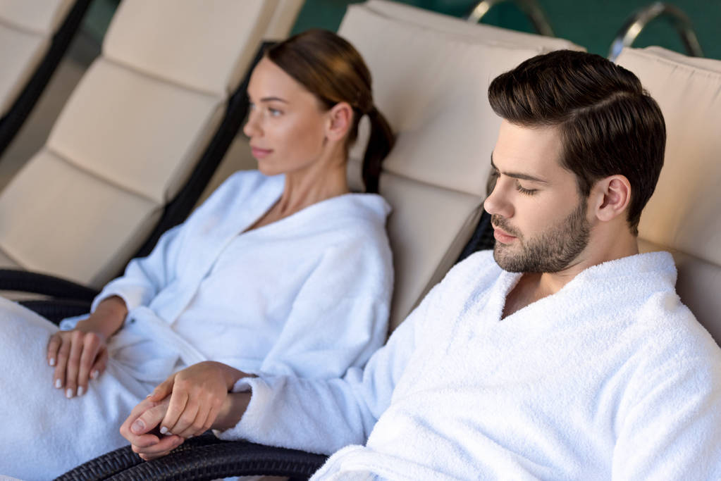 young couple in bathrobes holding hands while resting together in spa center - Photo, Image