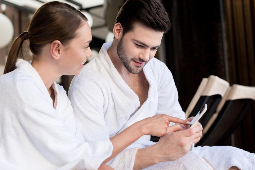 smiling young couple in bathrobes using smartphone together in spa center  - Photo, Image