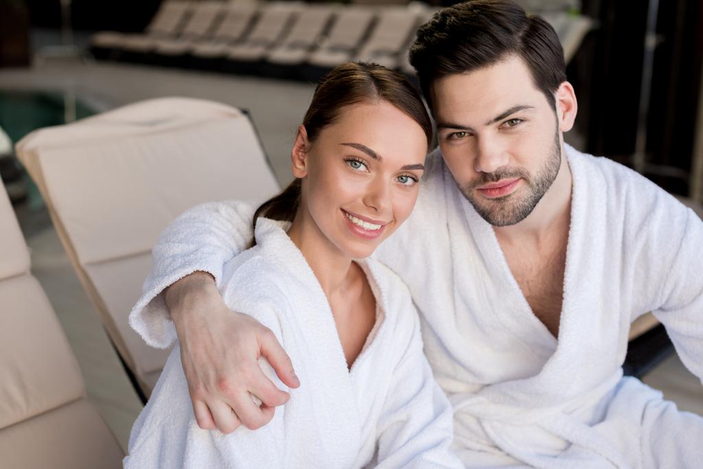 belo jovem casal em roupões de banho abraçando e sorrindo para a câmera no centro de spa
  - Foto, Imagem