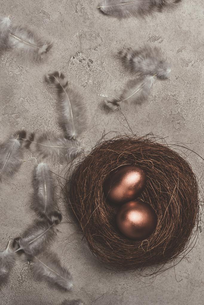 верхний вид золотых пасхальных яиц в гнезде с перьями на бетонной поверхности
 - Фото, изображение