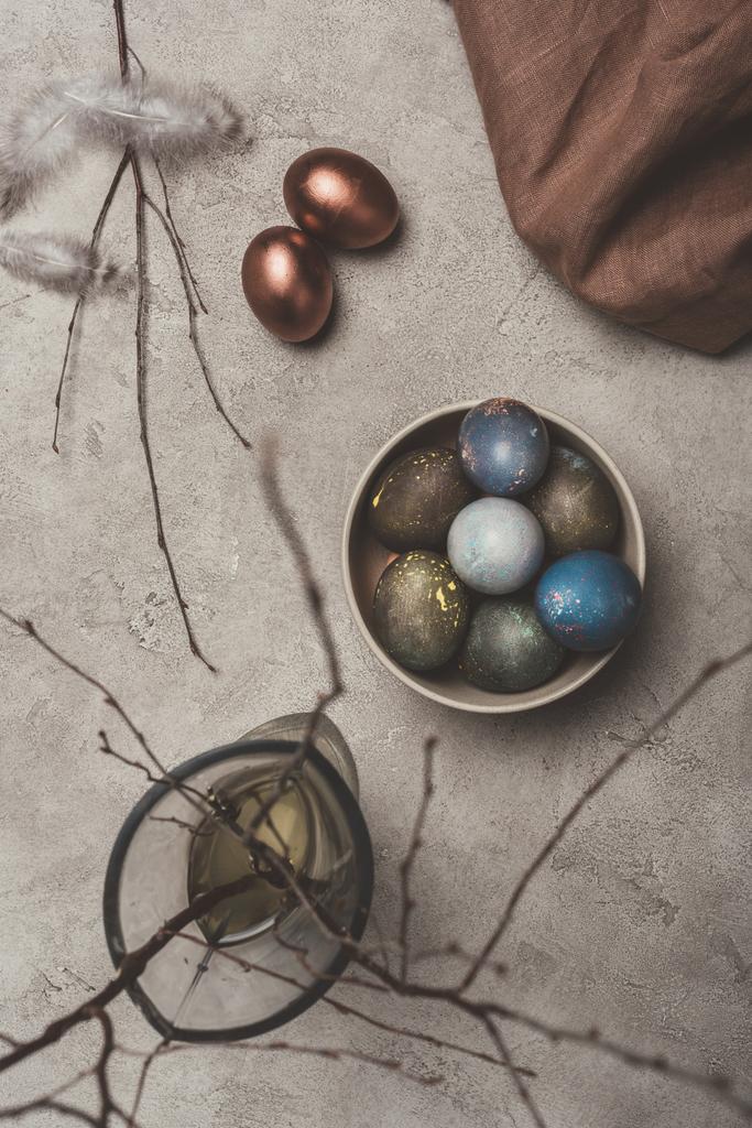 верхний вид окрашенных пасхальных яиц в чашу и ивовых ветвей в вазу на бетонную поверхность
 - Фото, изображение