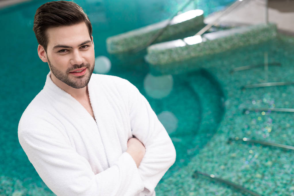 handsome young man in bathrobe standing with crossed arms and looking at camera in spa center - Photo, Image