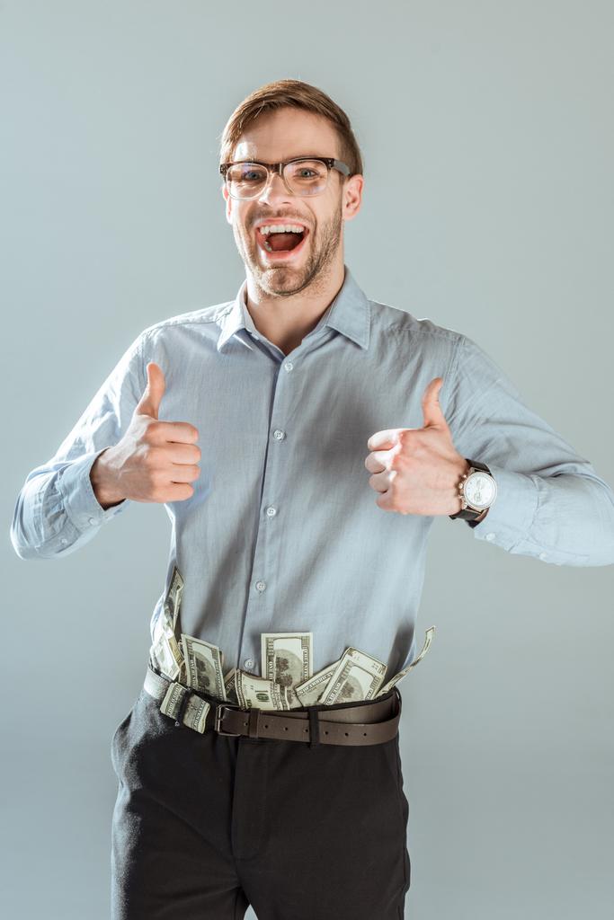 junger aufgeregter Geschäftsmann mit Dollarscheinen in der Hose, die vereinzelt Daumen hoch auf grau zeigt - Foto, Bild