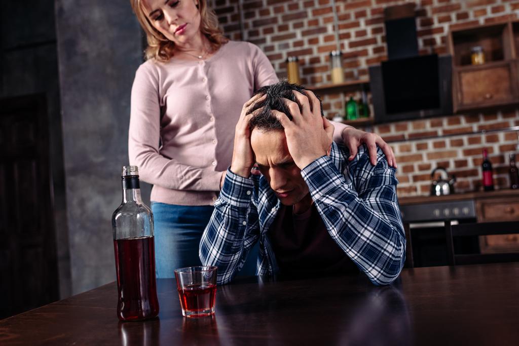 μεθυσμένος άντρας κάθεται στο τραπέζι, ενώ γυναίκα στέκεται κοντά στο σπίτι - Φωτογραφία, εικόνα
