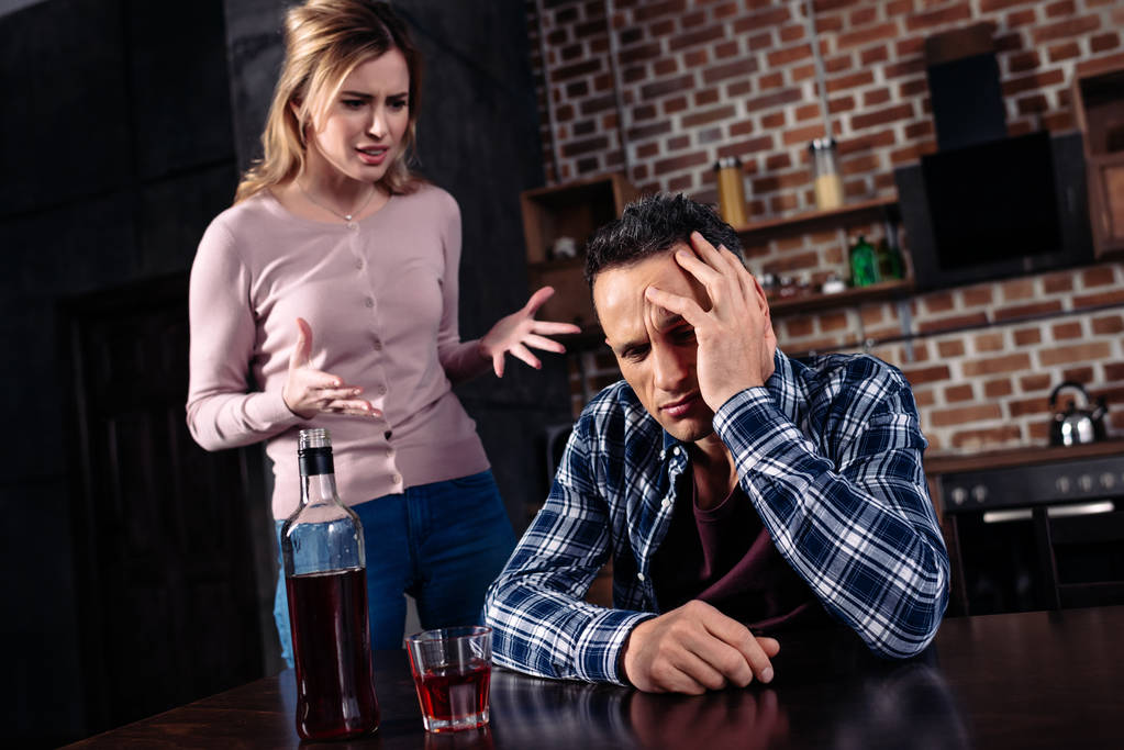 Betrunkener sitzt am Tisch, während streitende Ehefrau in der Nähe zu Hause steht - Foto, Bild