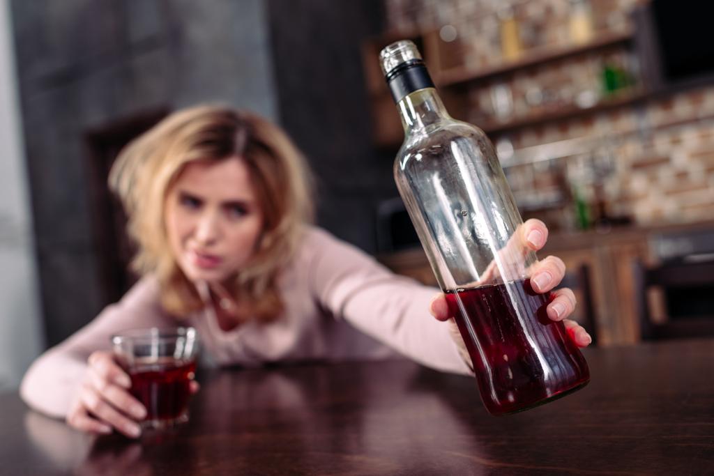 вибірковий фокус п'яної жінки, що приймає пляшку алкоголю, сидячи за столом вдома
 - Фото, зображення