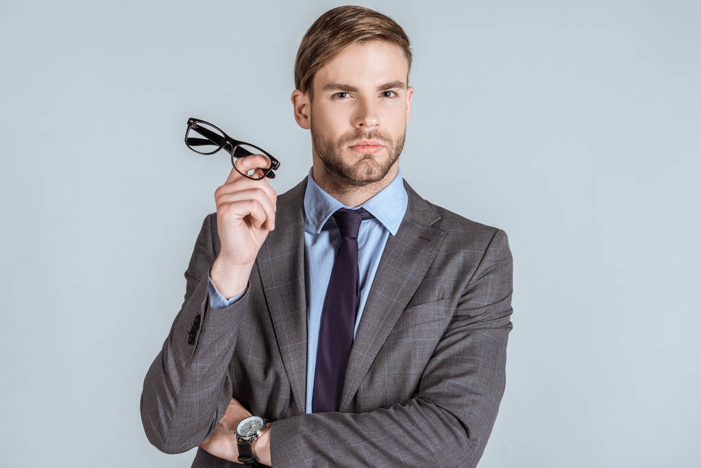 Young confident businessman holding glasses isolated on grey - Photo, Image